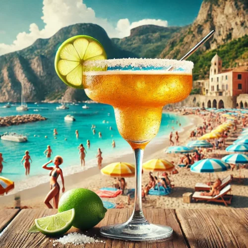Amber margarita with a salted rim, lime wheel and straw on a wooden table above a busy beach with striped umbrellas, a rocky headland and colourful buildings
